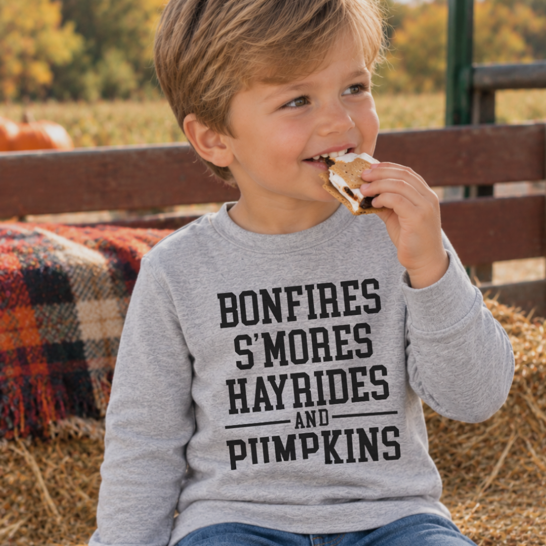 Young boy eating a s’more while wearing the Fall Traditions T-shirt featuring a bonfires, s’mores, hayrides and pumpkins graphic.