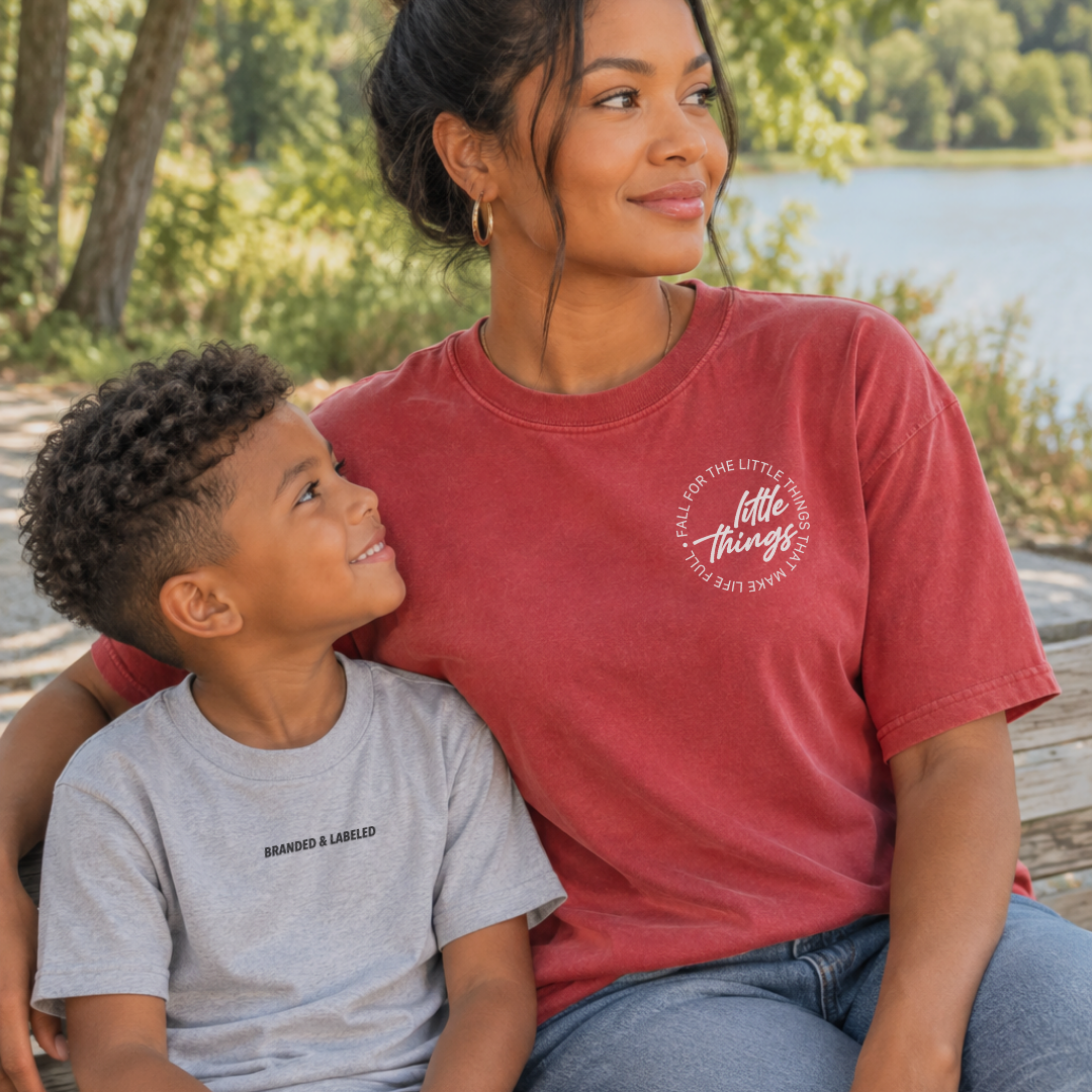 Woman and young boy sitting together on a bench. The mother wears a crimson colored tee featuring the “Fall for the Little Things” design, while the boy wears a heather light gray tee featuring the Branded & Labeled design both from Branded & Labeled