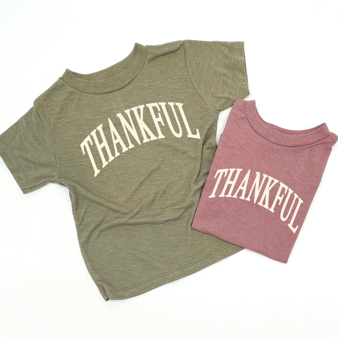 Two kids’ T-shirts in olive and mauve, both featuring a large “thankful” graphic across the front.