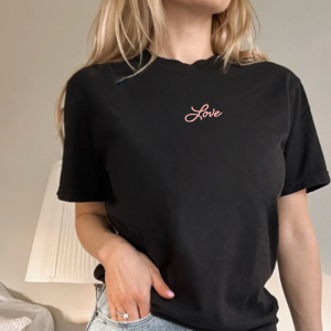 woman standing, wearing a black short sleeve tshirt with the word "Love" writen in a cursive script font in pink ink printed small on the front just below the collar