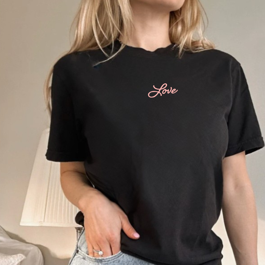 woman standing, wearing a black short sleeve tshirt with the word "Love" writen in a cursive script font in pink ink printed small on the front just below the collar