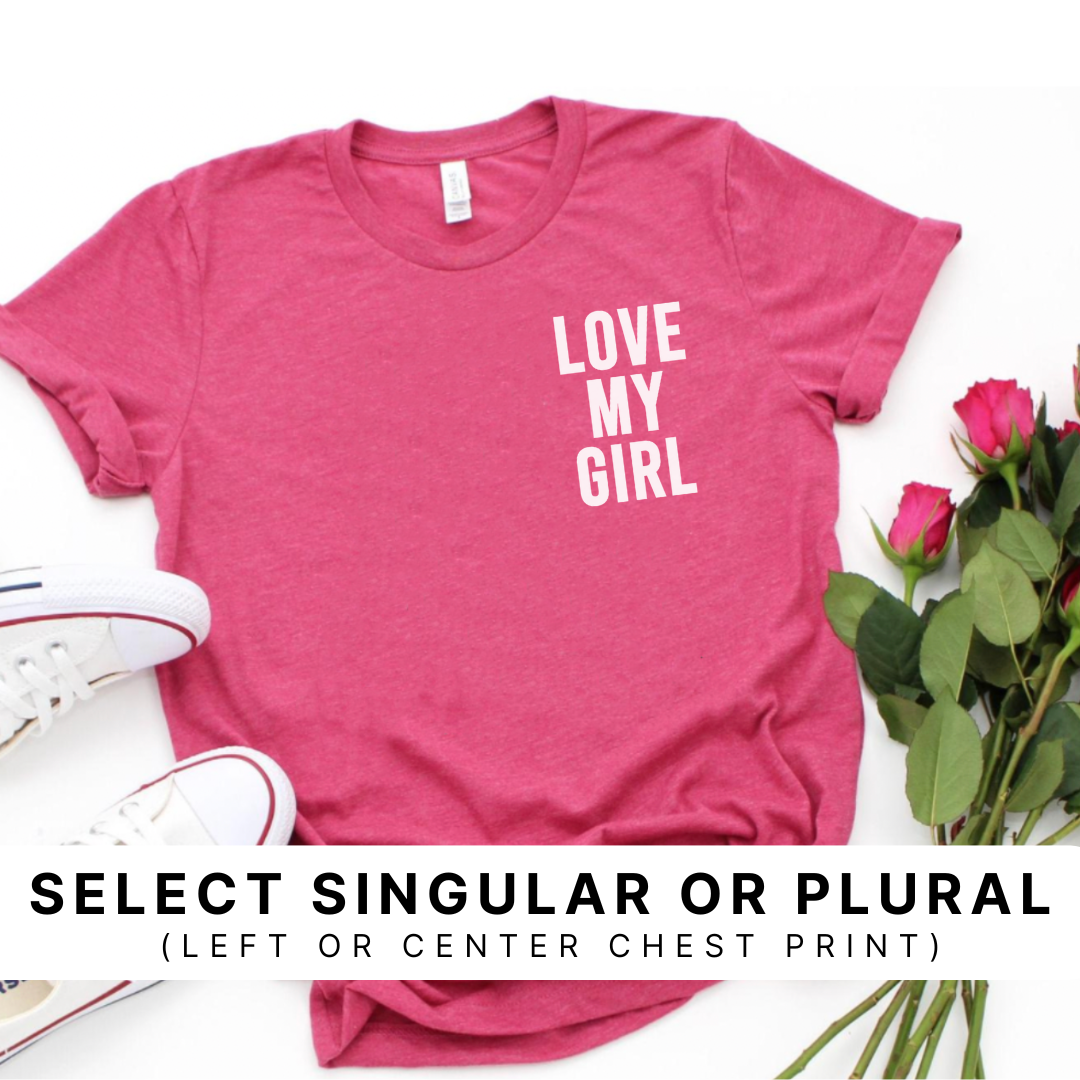 Flat lay of a heather raspberry graphic tee featuring the “Love My Girl” design in white ink printed on the left chest, styled with flowers around the shirt with a pair of white shoes.