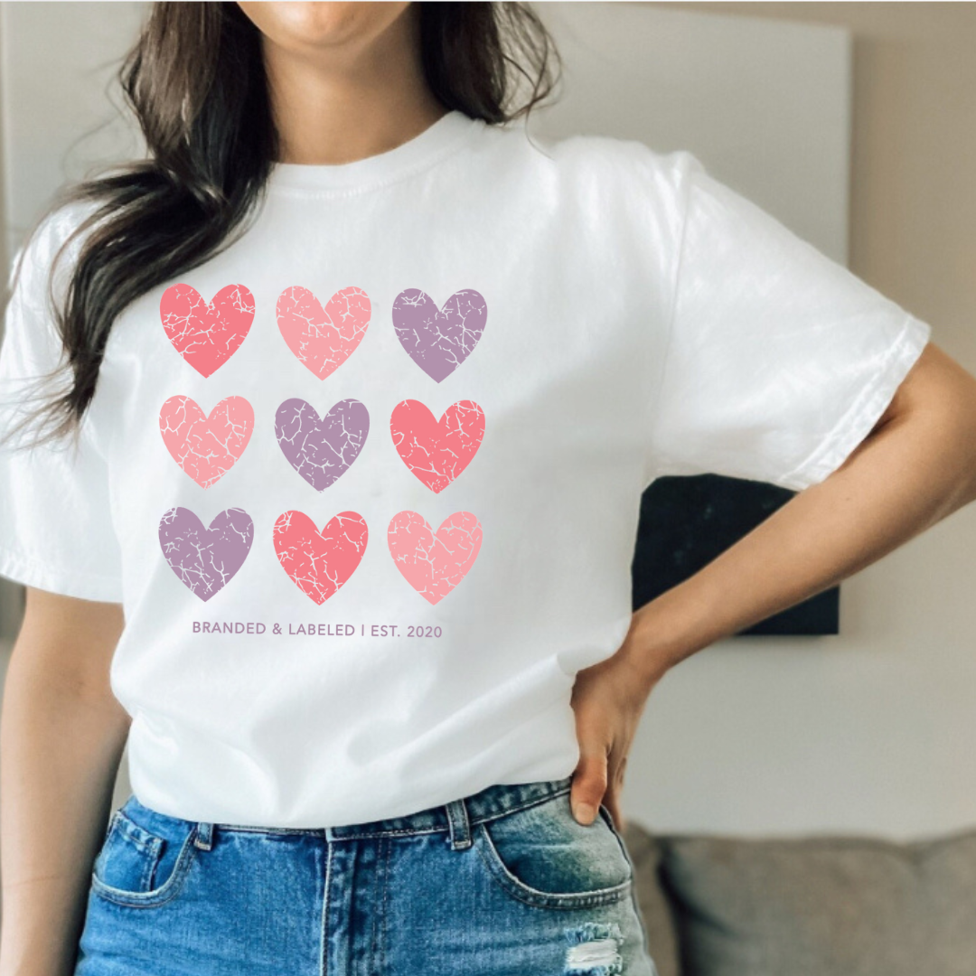 Close-up lifestyle photo of a woman wearing the “Loved Over Time” everyday tee, tucked in, featuring distressed hearts in light pink, purple, and dark pink tones on the front.