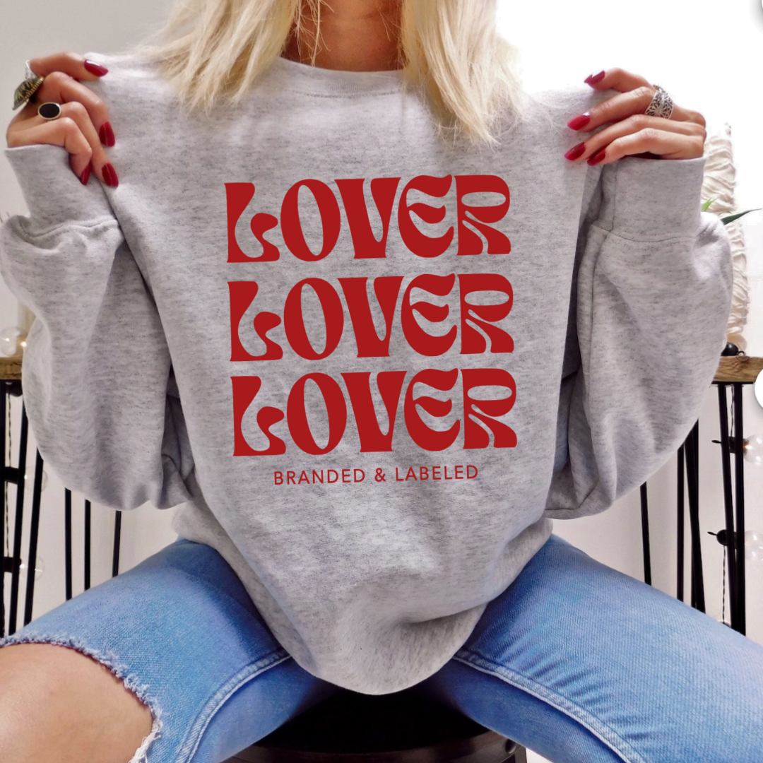 lady sitting, modeling the heather light gray fleece pullover with "Lover" repeated 3 times stacked on top of each other and "Branded & Labeled" printed in a smaller font size all in red ink.
