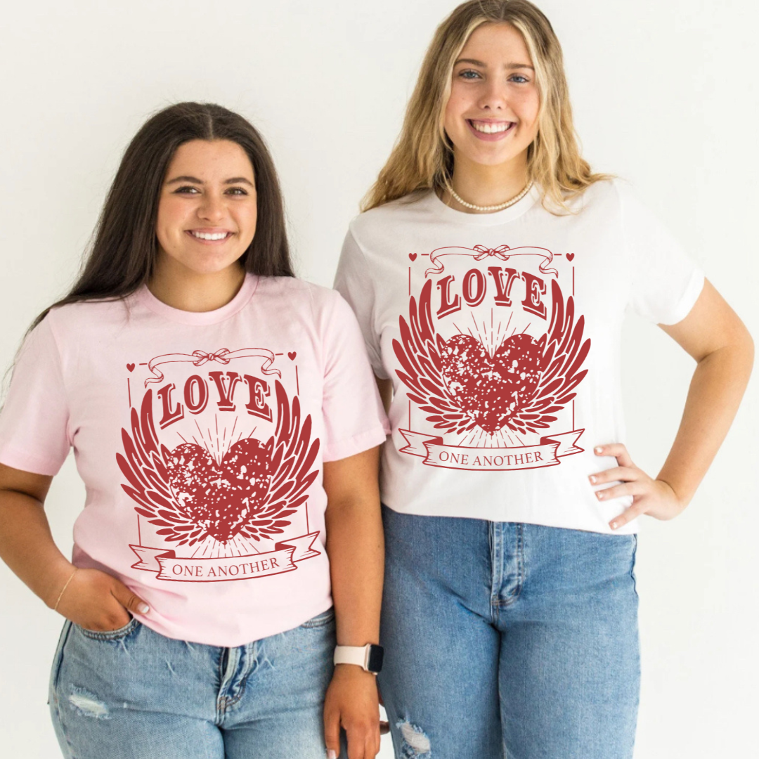 2 women standing side by side wearing the Love One Another graphic design, one wearing a white shirt and the other wearing a pink shirt