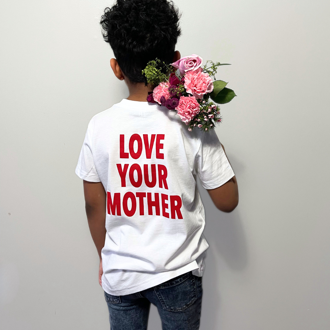 Boy standing with his back facing the camera, modeling a Branded & Labeled graphic tee with “Love Your Mother” printed in bold red lettering while holding a small bouquet of flowers.