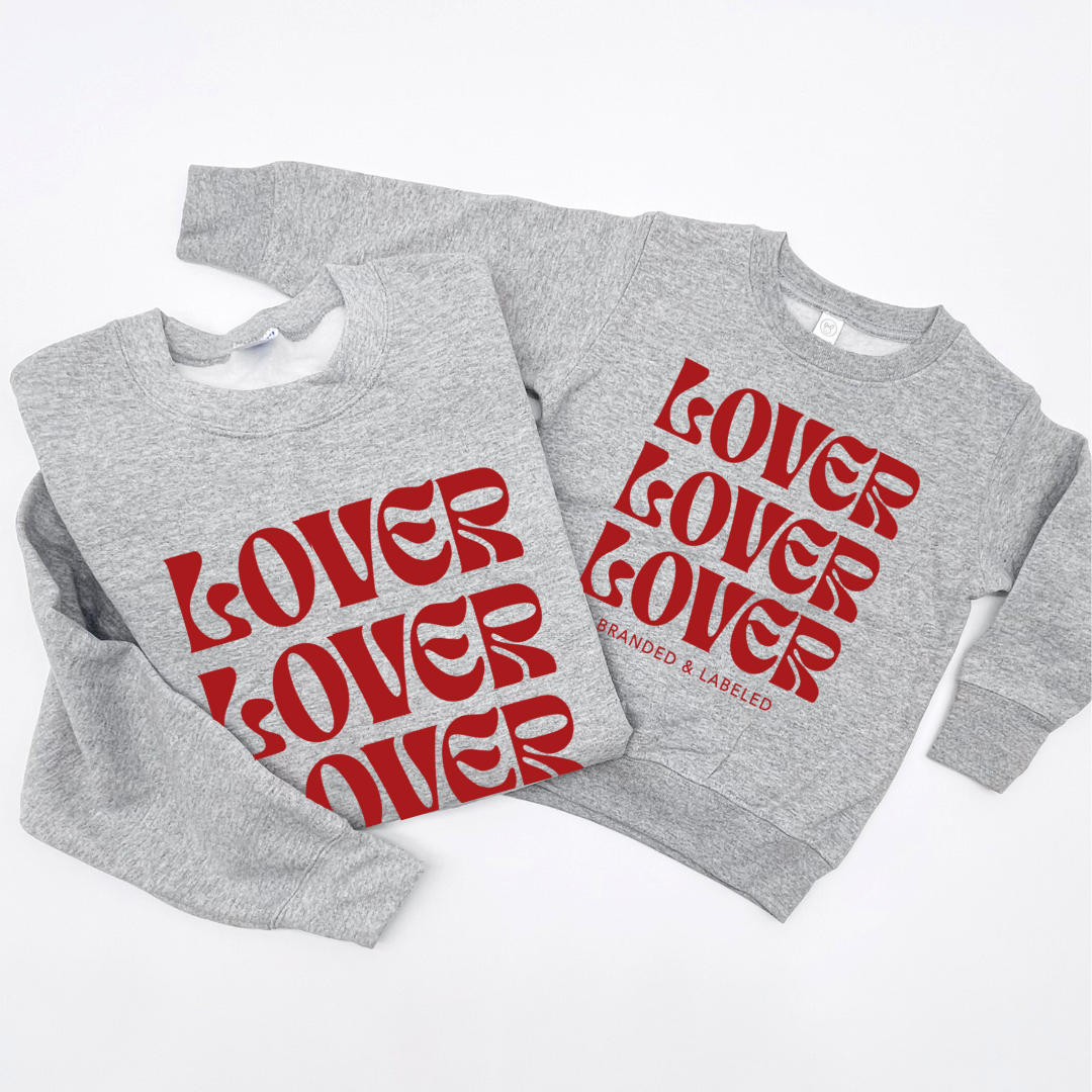 Flat lay image showing an adult and kids heather light gray fleece pullover placed side by side, both featuring the “Lover on Repeat” graphic on the front.