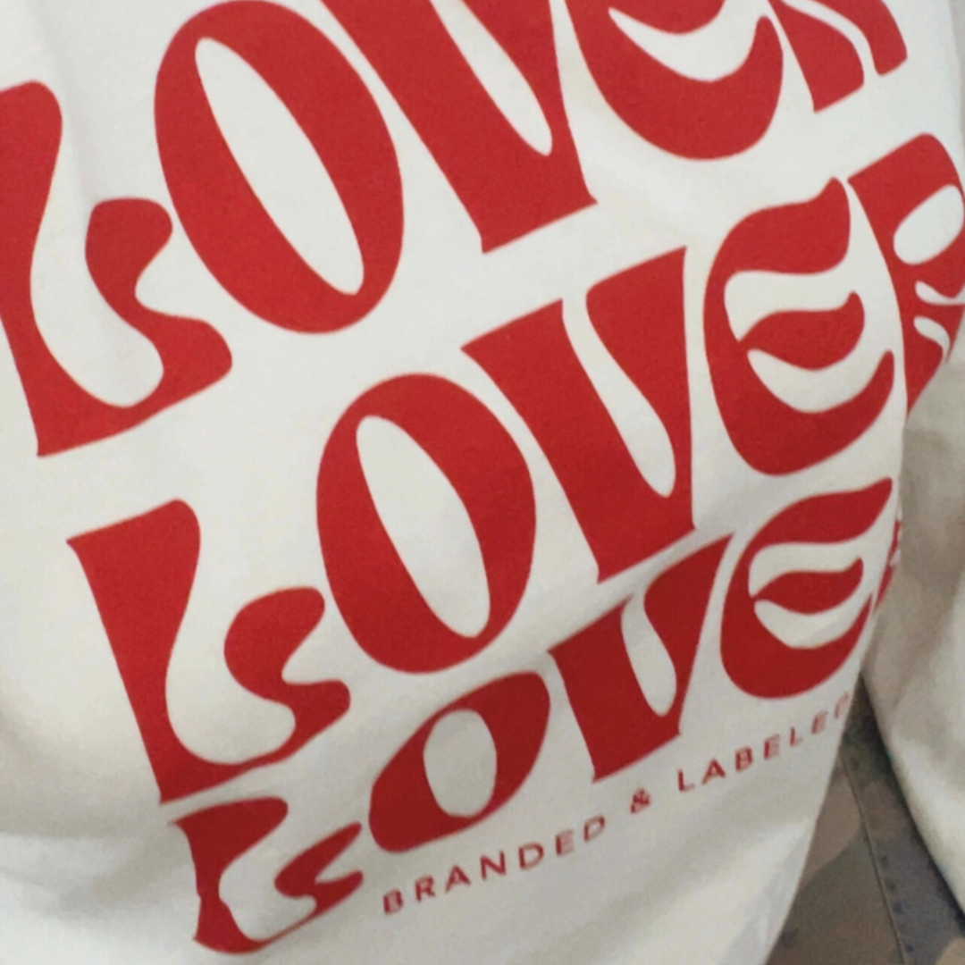 Model wearing the Branded & Labeled “Lover On Repeat” graphic long sleeve in ivory, featuring the word “Lover” printed three times in bold red ink with subtle Branded & Labeled branding underneath.