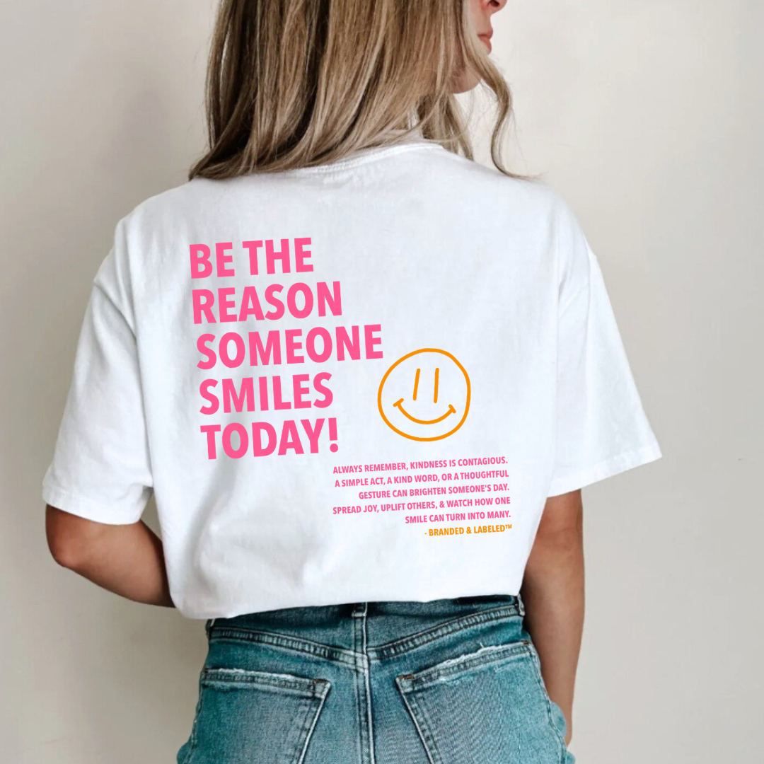 Woman wearing a graphic tee featuring the back design “Be the Reason Someone Smiles Today” with a kindness message and Branded & Labeled signature.