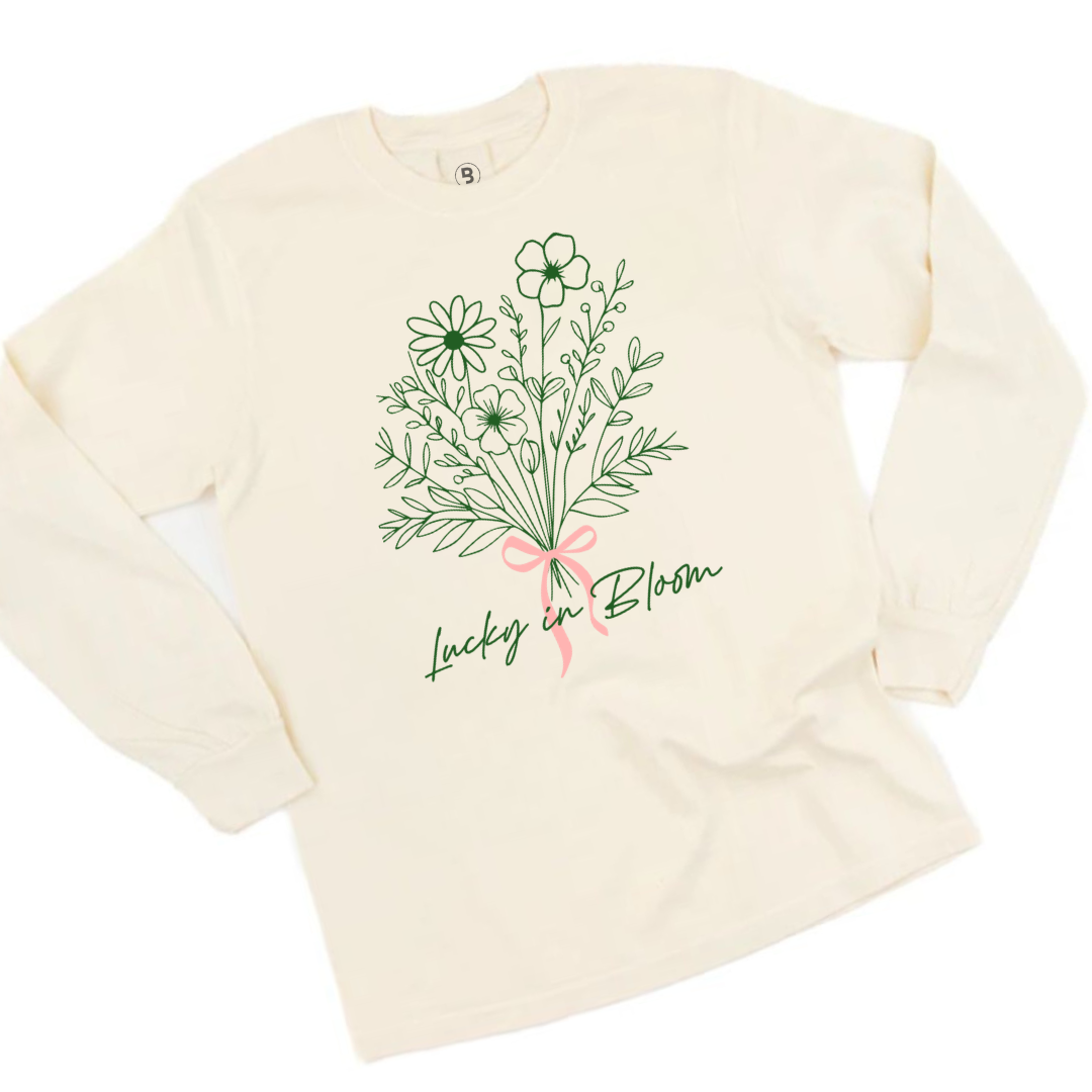 Adult ivory colored premium long sleeve with a floral bouquet in green , tied with a pink bow, with text that reads "Lucky In Bloom" beneath it