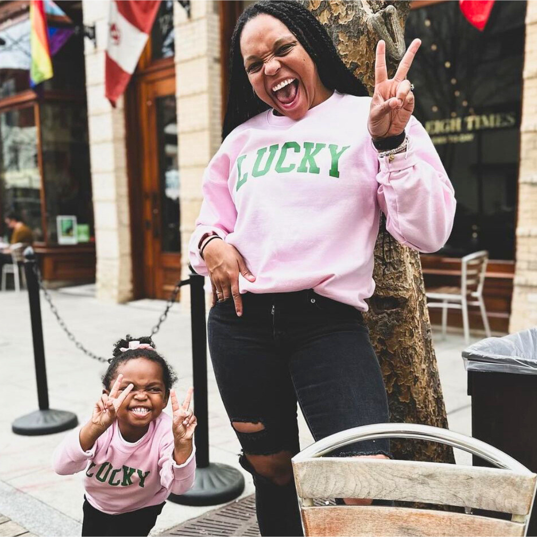 Mom and daughter wearing matching pink fleece pullovers with ‘LUCKY’ printed in bold green varsity lettering, standing outdoors.