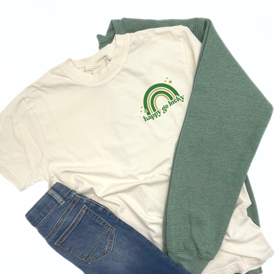 Adult ivory colored short sleeve tee styled wiht a green fleece pullover and denim jeans with a green and gold rainbow with text that reads "happy go lucky" and printed on the let chest of the shirt