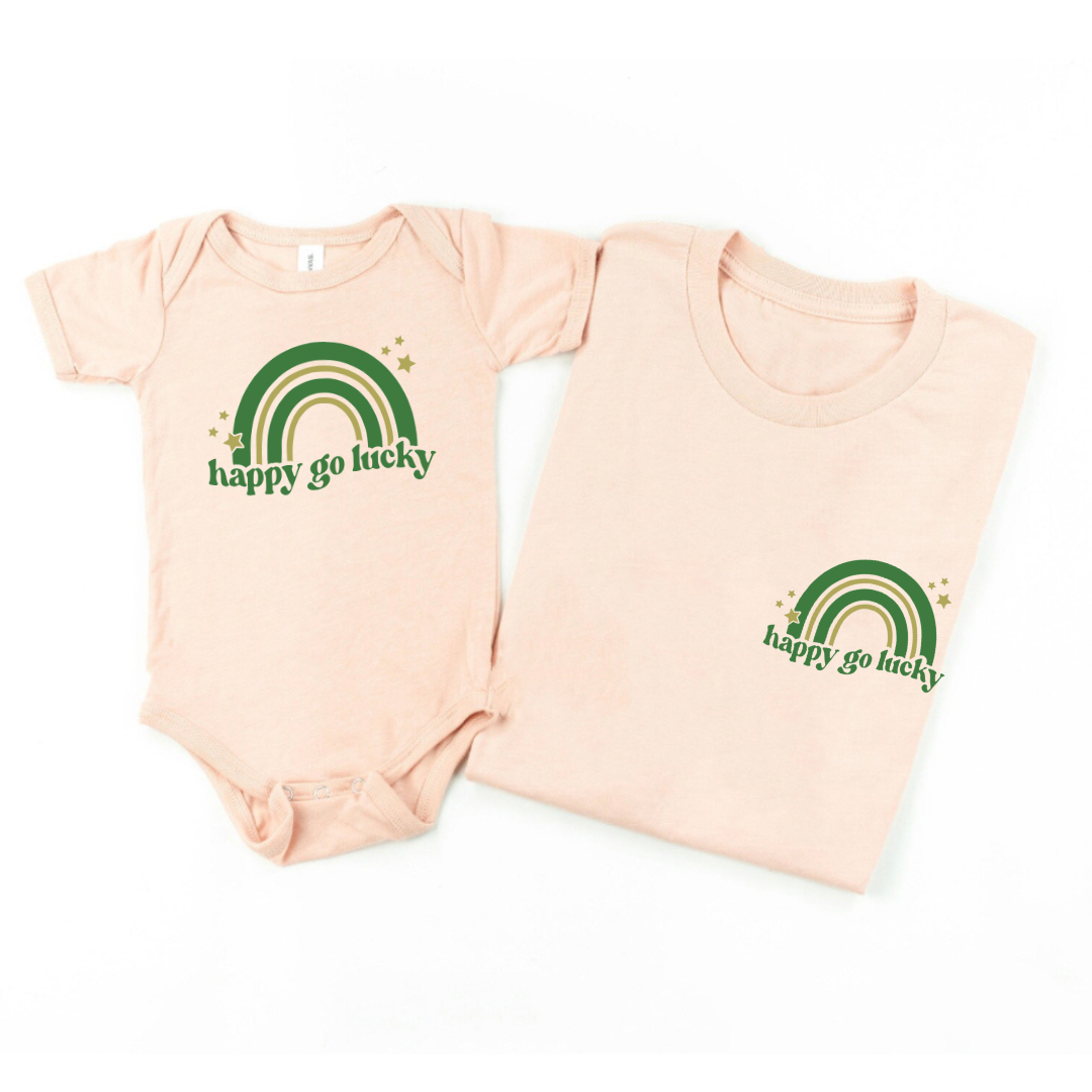 MATCHING SET OF 2 - ONE ADULT TEE FOLDED AND ONE CHILD BODYSUIT - BOTH LAYING SIDE BY SIDE IN THE COLOR PEACH WITH THE "HAPPY GO LUCKY" DESIGN THAT FEATURES A GREEN AND GOLD COLORED RAINBOW WITH HAPPY GO LUCKY PRINTED UNDERNEATH