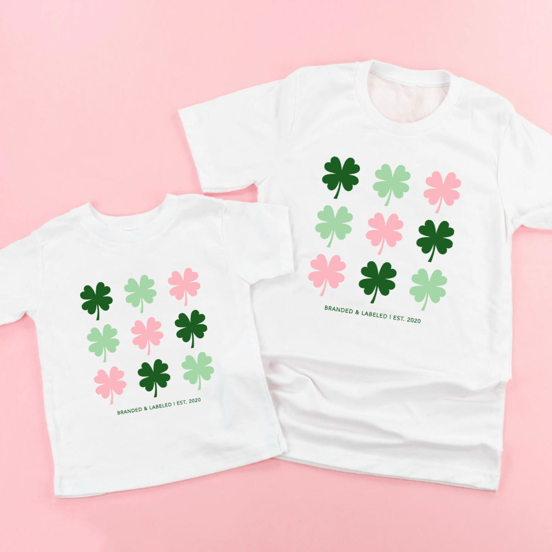 Matching set of 2 white tees - one adult and one kid with a total of 9 shamrocks all alternating colors between dark green, light green, and pink with the text "Branded & Labeled | Est. 2020" underneath them