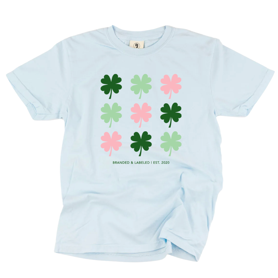 Adult light blue short sleeve tee with a total of 9 shamrocks all alternating colors between dark green, light green, and pink with the text "Branded & Labeled | Est. 2020" underneath them