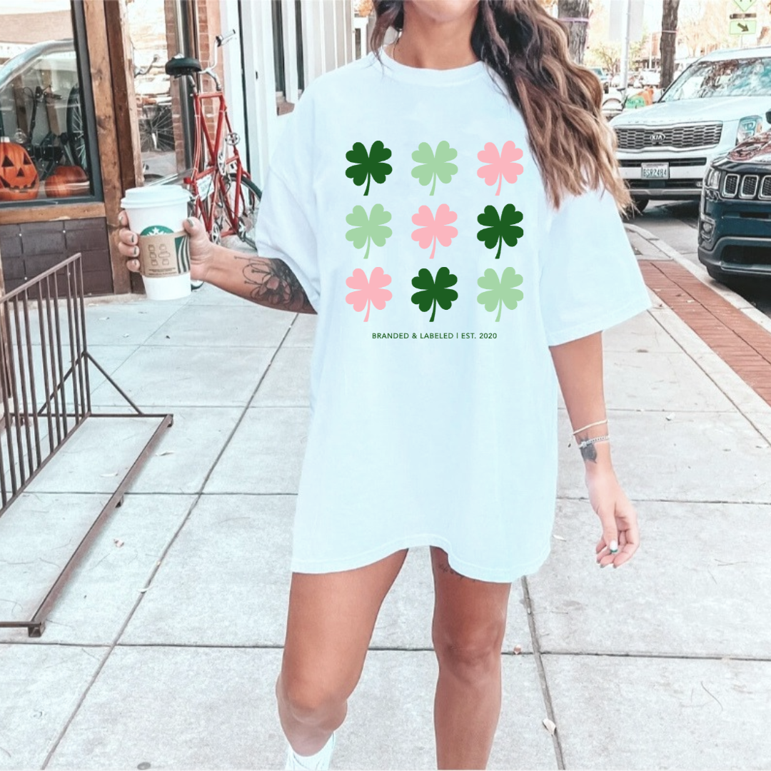 WOMAN STANDING OUTSIDE HOLDING A COFFEE WHILE MODELING AN OVERSIZED WHITE TEE WITH A SERIES OF 9 SHAMROCKS ALL ALTERNATING COLORS BETWEEN DARK GREEN LIGHT GREEN, AND PINK WITH TEXT THAT READS "BRANDED & LABELED | EST. 2020"