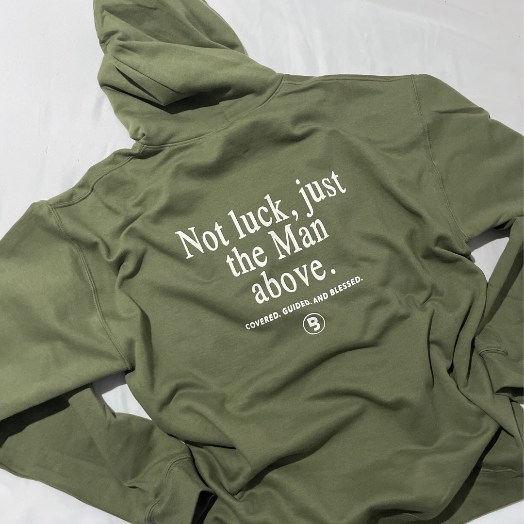 Flat lay image of the back of a youth military green hoded pullover with the "Man Above" graphic that has "Not luck, just the man above: Covered. Guided. And Blessed" followed by the signature Branded & Labeled "B'" logo