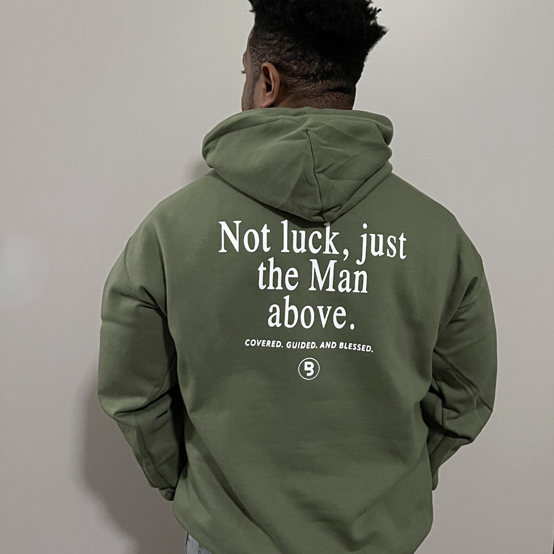 Man photographed from behind modeling a military green hooded pullover with the "Man Above" graphic design which has text that reads "Not luck, just the man above" followed by "Covered. Guided. And Blessed" with the signature "B" branded & Labeled logo