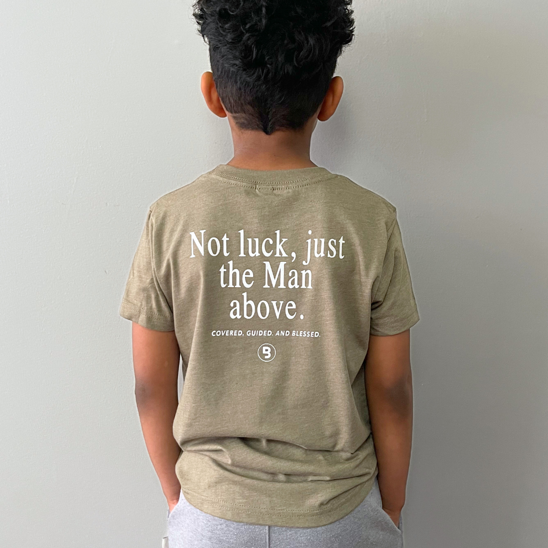 young boy photographed from behind while modeling the "Man Above" graphic tee which shows the text "Not luck, just the Man above - Covered. Guided, And Blessed" with the signature Branded & Labeled "B" logo
