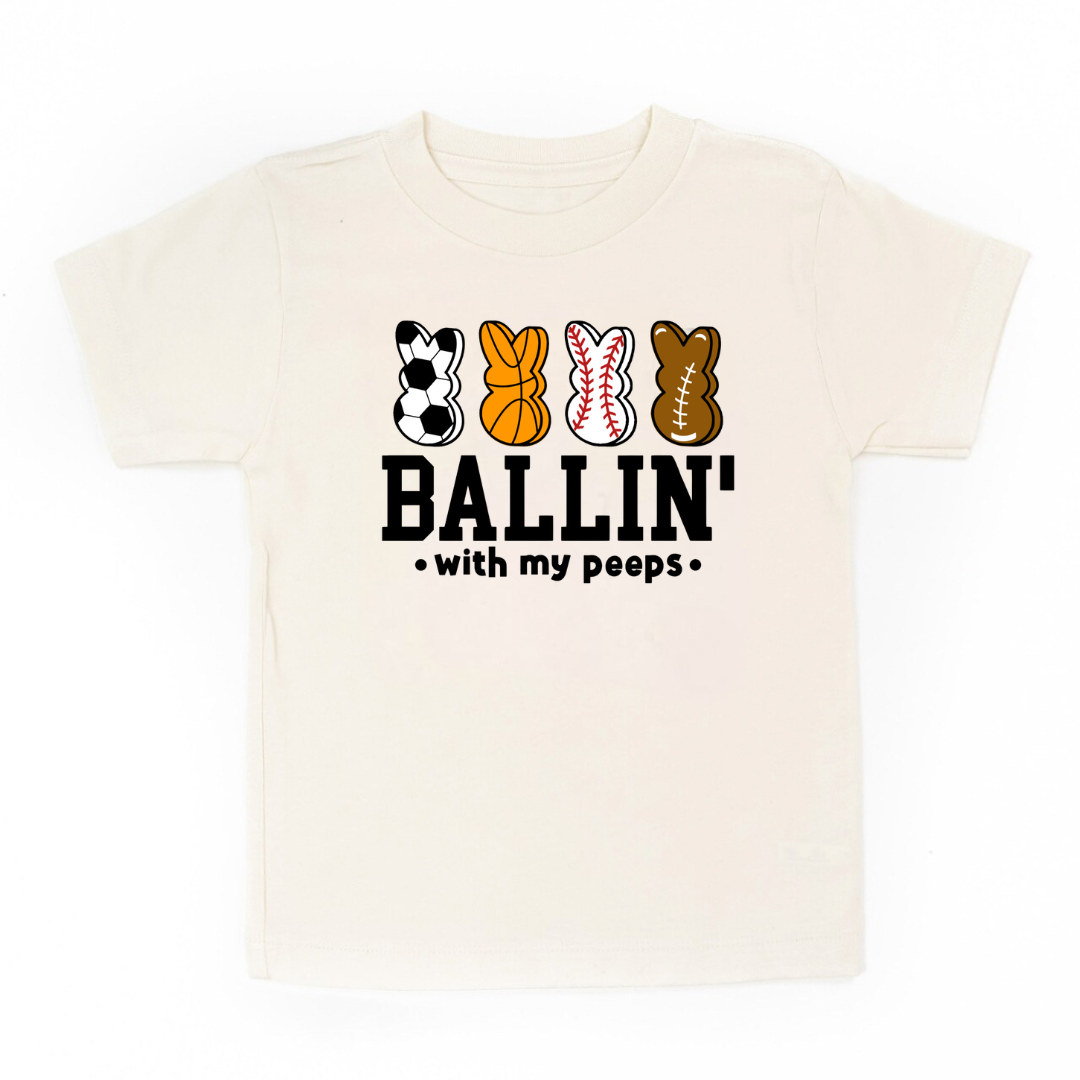 Kids graphic tee featuring bunny shapes designed as different sports balls with the phrase “Ballin With My Peeps” from Branded & Labeled.