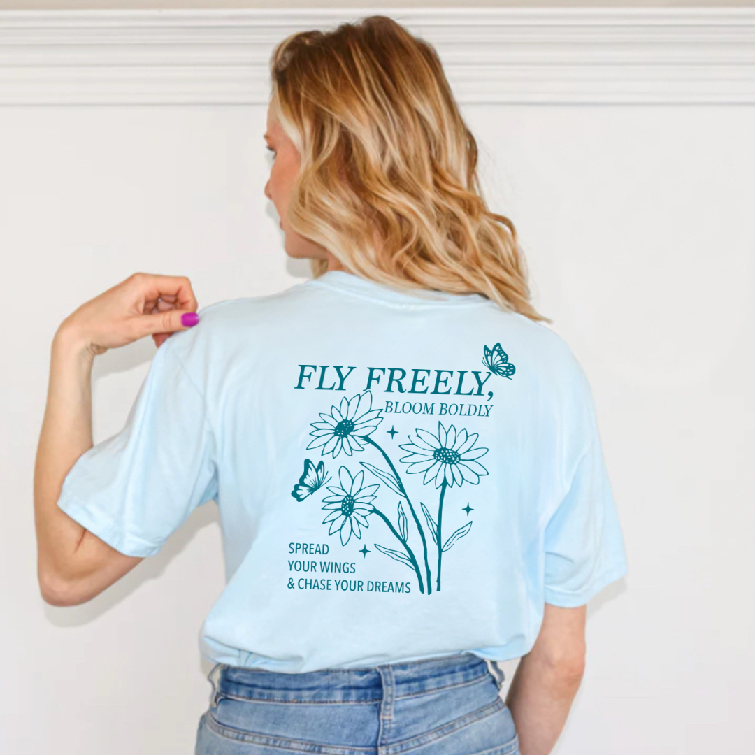 Woman modeling a graphic tee showing the back design that reads “Fly Freely, Bloom Boldly. Spread Your Wings and Chase Your Dreams” from the Branded & Labeled spring collection.