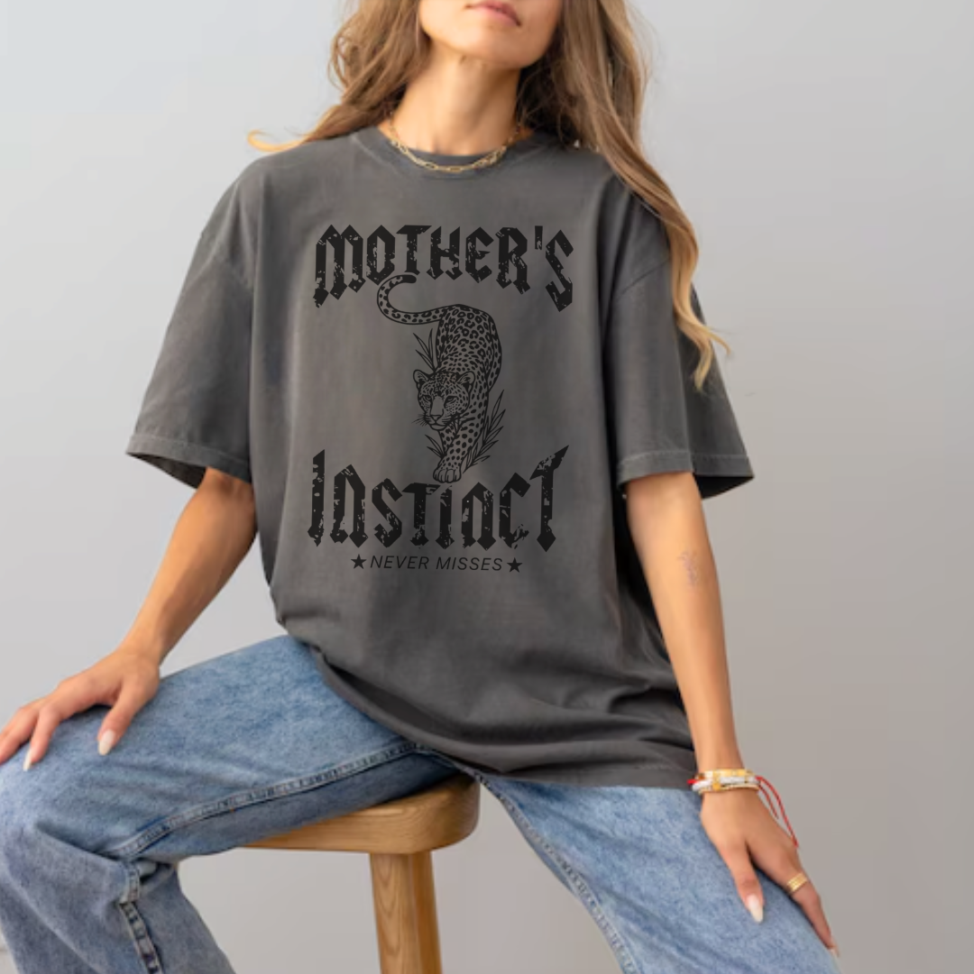 Woman wearing the “Instinct Of A Mother” premium graphic tee from Branded & Labeled featuring a bold cheetah design, and text that reads "Mother's instinct: Never Misses" posing confidently on a stool