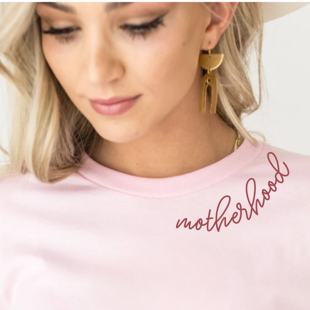 Close-up of a woman wearing an adult premium tee, showing “motherhood” in a cursive font curved around the left side of the collar from Branded & Labeled