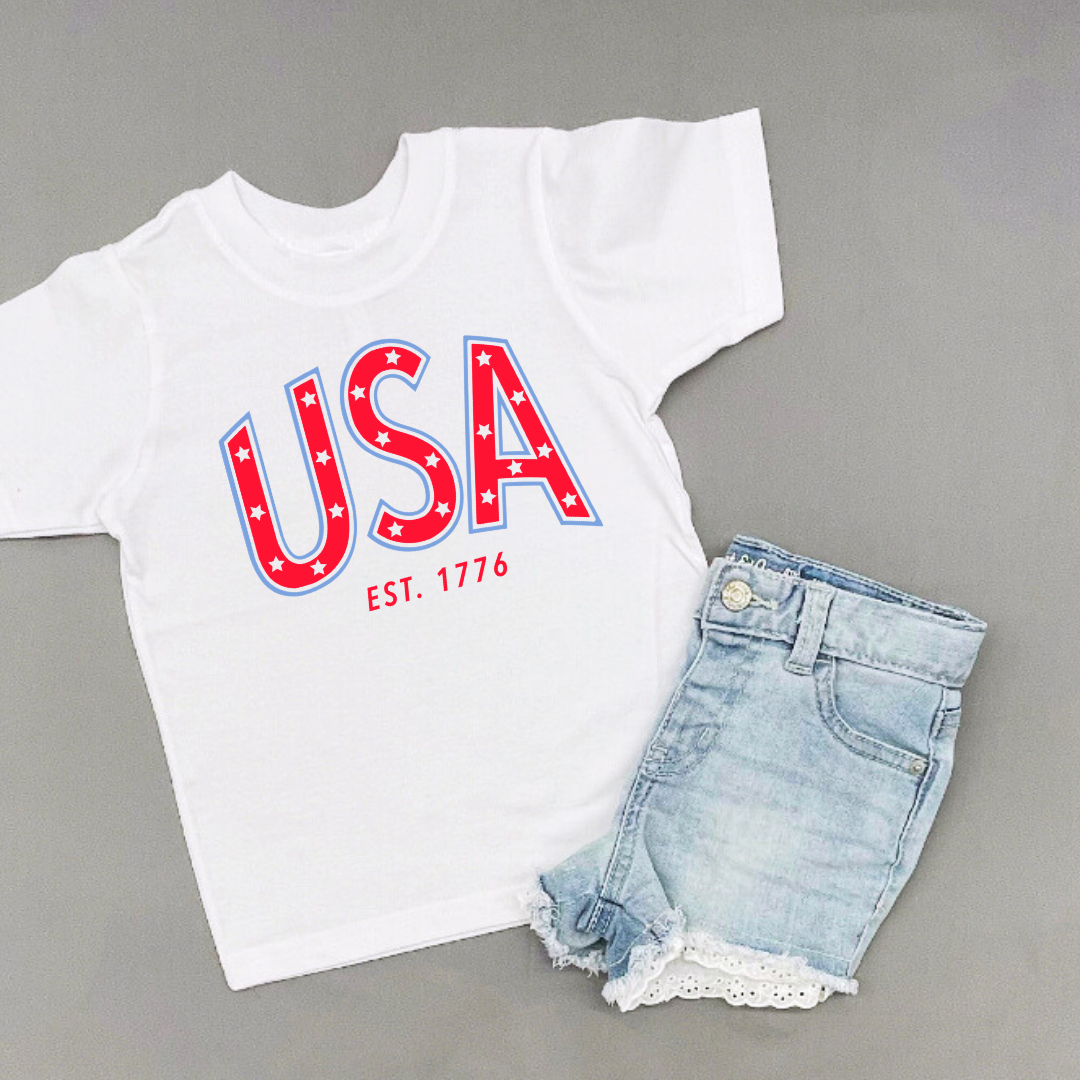 Kids graphic tee featuring bold “USA” typography with est. 1776 detail in red and blue patriotic colors, part of the American Classics Collection by Branded & Labeled.