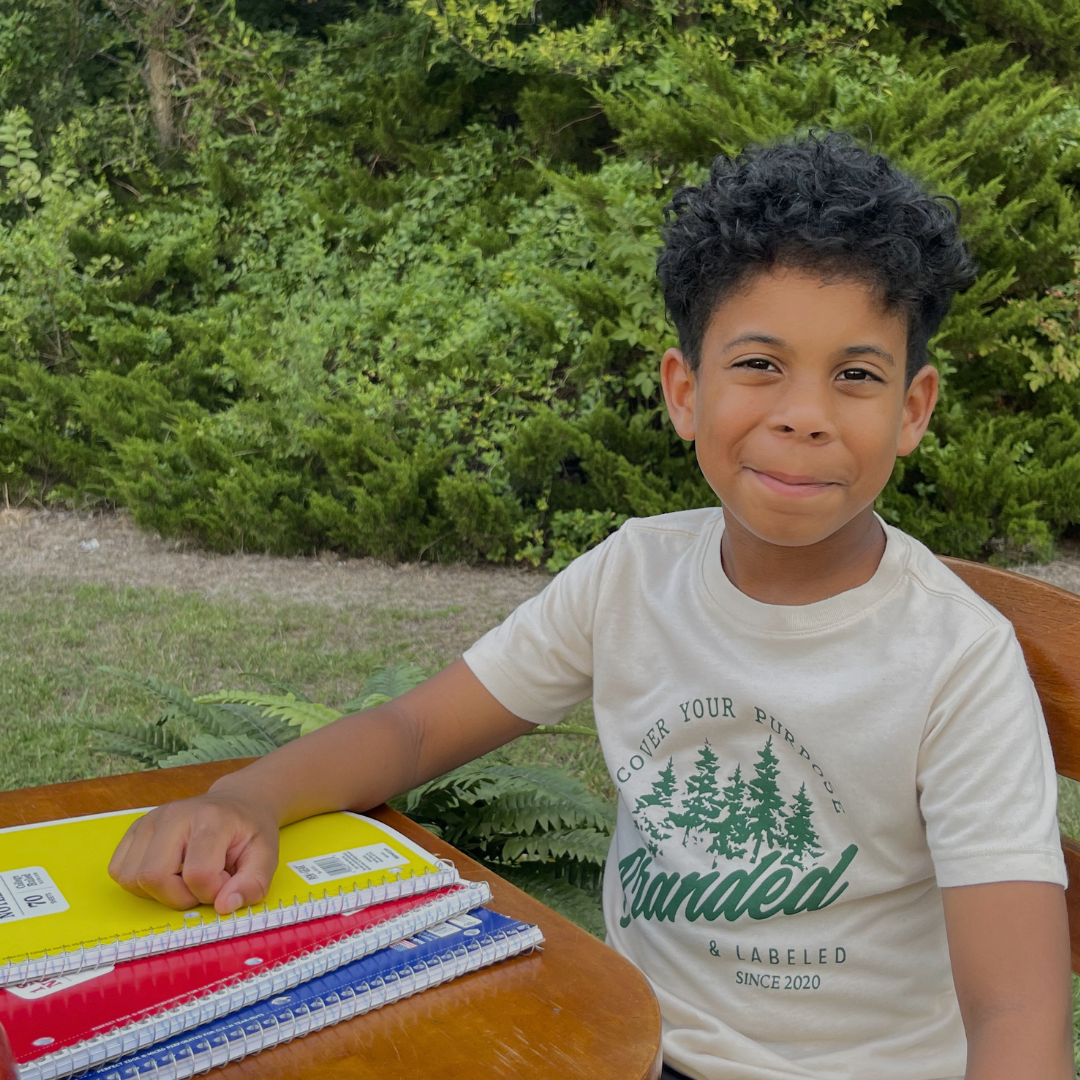 Young boy wearing a Discover Your Purpose kids graphic t-shirt while sitting at a school desk, featuring an inspirational design from Branded & Labeled's The Next Chapter collection.