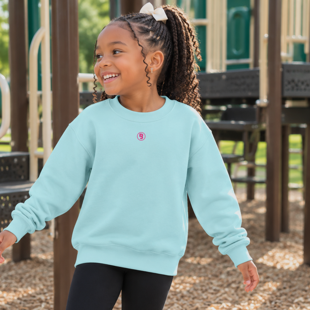 Young girl wearing the Signature Dream Cloud Youth Premium Fleece Pullover from Branded & Labeled in a soft chill blue color with a subtle hot pink signature logo near the collar while playing on a playground. The cozy youth fleece sweatshirt is shown as a comfortable everyday layer for school, outdoor play, and family adventures.
