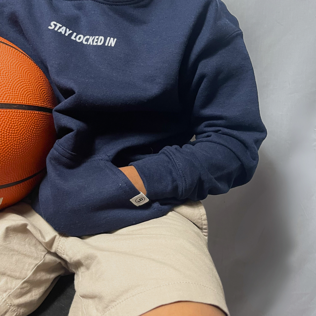 Young boy wearing the Stay Locked In youth premium hooded pullover while holding a basketball, showcasing the “Stay Locked In” message near the front collar and the signature sewn “B” logo on the kangaroo pocket.
