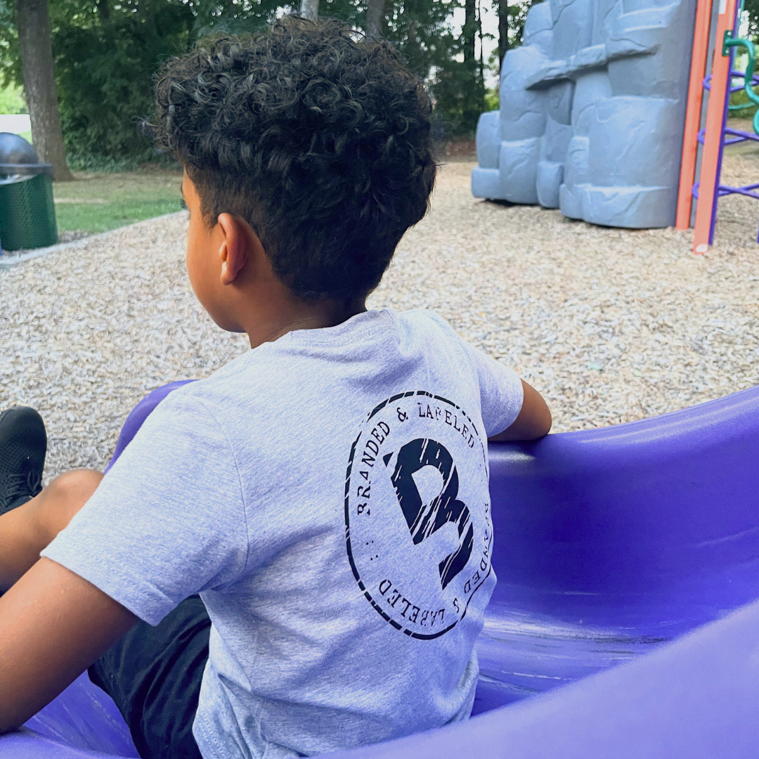 Young boy wearing the Branded & Labeled Origins kids graphic tee while sliding down a playground slide, featuring a large distressed circular B logo on the back.