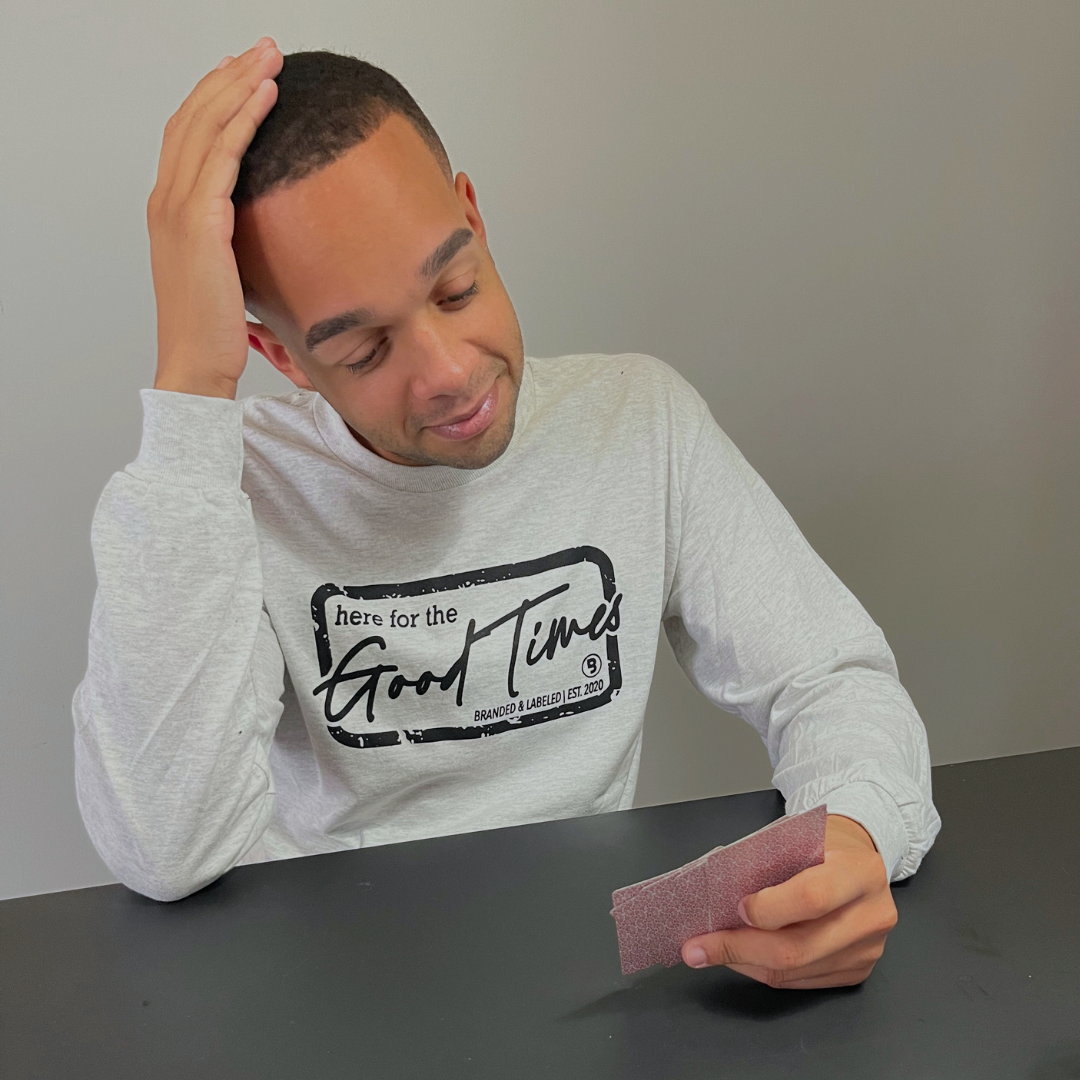 Man wearing the Good Times Core Long Sleeve in light ash gray while sitting at a table playing cards, featuring a Here for the Good Times graphic on the front from Branded & Labeled