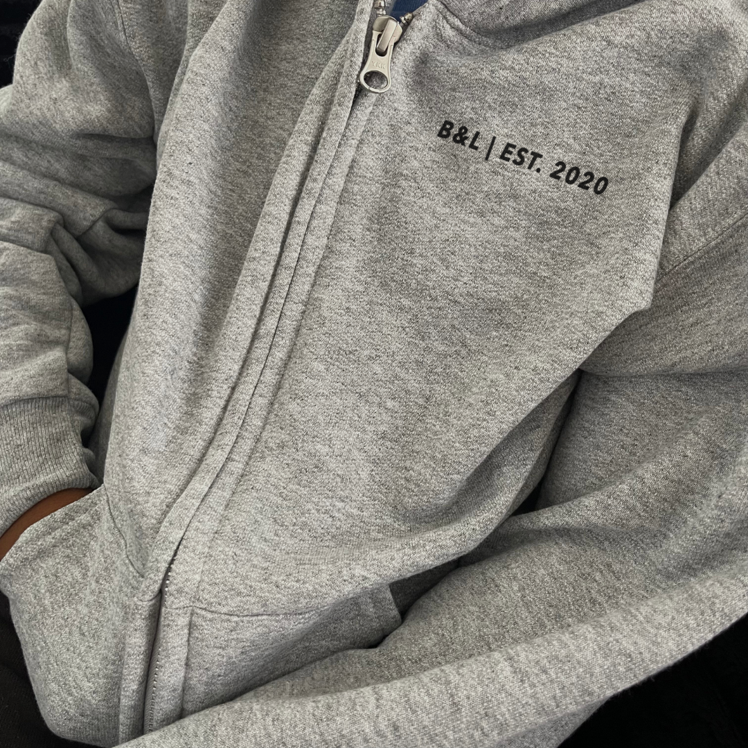 Boy shown from the front wearing the Riverstone Youth Premium Full Zip hoodie with B&L EST. 2020 printed on the front left chest and a signature B logo on the back from Branded & Labeled