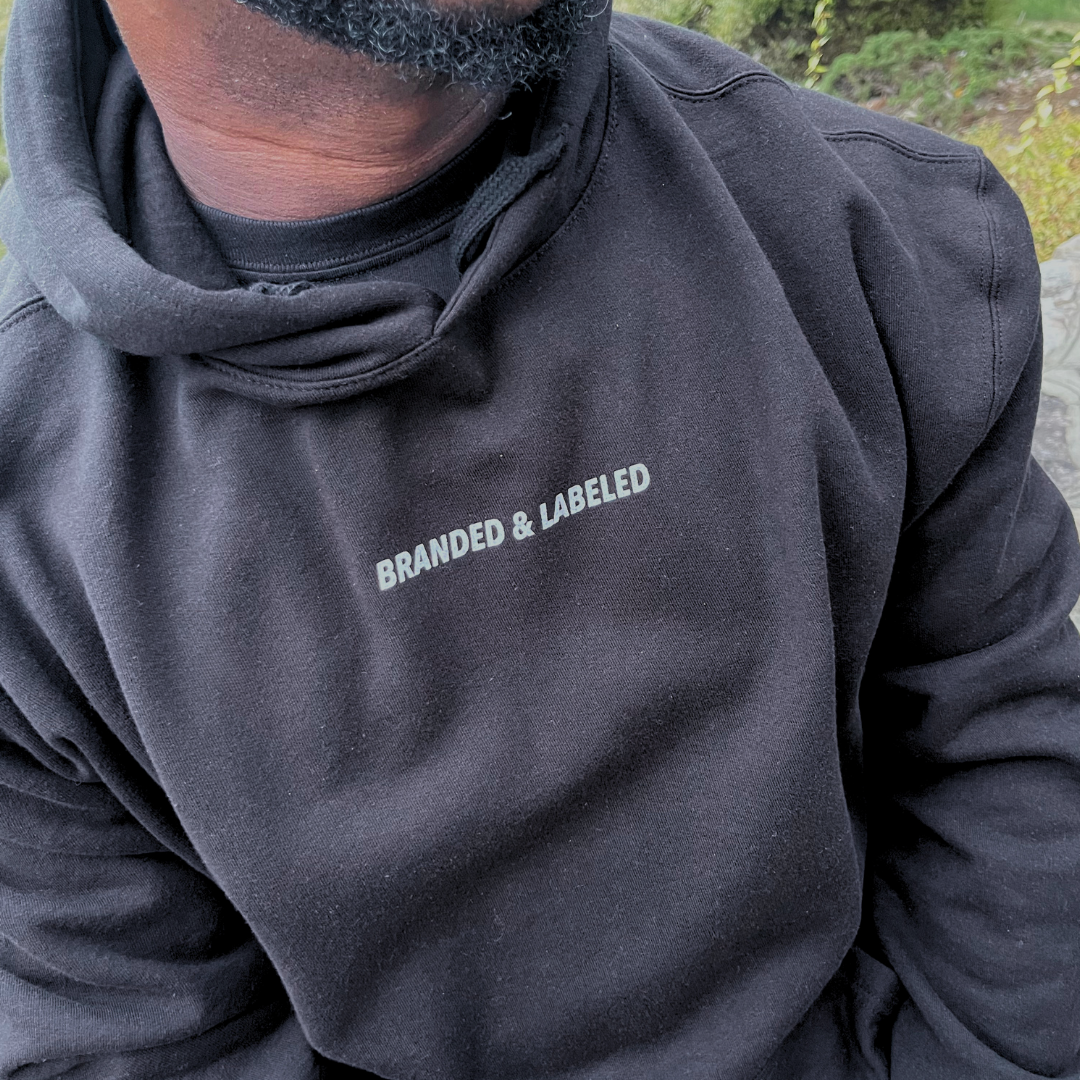 Man wearing The Label Adult Everyday Hooded Pullover in black, featuring subtle Branded & Labeled lettering printed in white near the collar.